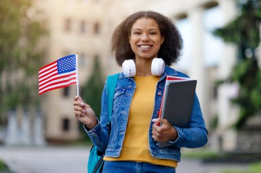Yurtdışında oku. Gülümseyen siyah kız öğrenci, Amerikan bayrağıyla poz veriyor, Sırt çantası ve çalışma kitapları olan mutlu Afrikalı genç Amerikalı kadın Üniversite binasının yanında duruyor, Kopya Boşluğu