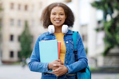 Pozitif siyah bayan öğrenci sırt çantası ve çalışma kitaplarıyla üniversite kampüsünde poz veriyor, kulaklık takıyor ve kameraya gülümsüyor.