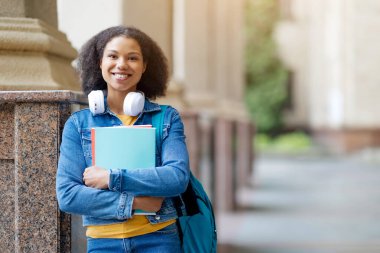 Siyahi Afrikalı Amerikalı kız öğrenci üniversite koridorunda ayakta duruyor, kitaplarını tutan ve boynuna beyaz kulaklık takan mutlu genç bir kadın, kameraya bakıyor, boşluğu kopyalıyor.