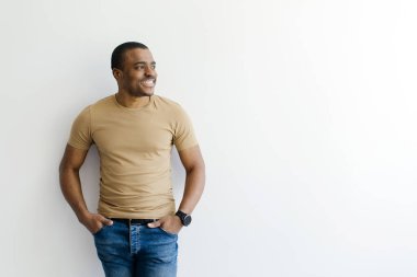 Beyaz stüdyo duvarına yaslanmış bej tişörtlü ve mavi kotlu Afro-Amerikan bir adam gülümseyen, genç, siyah bir erkek ellerini cebinde tutuyor ve fotokopi odasına bakıyor.