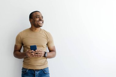 Bej tişörtlü, gülümseyen ve iki eliyle akıllı bir telefon tutan Afro-Amerikan bir adam. Mutlu siyah erkek stüdyo ortamında düşünceli bir şekilde bakıyor.