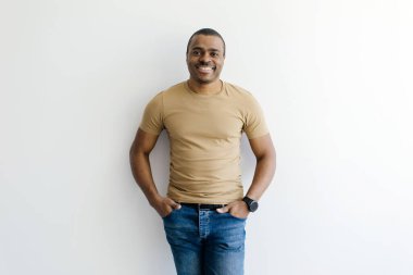 Gülümseyen, bej tişört ve kot pantolon giyen, elleri ceplerinde beyaz bir stüdyonun arka planında duran, kendine güvenen siyah bir erkek kameraya bakıyor, boşluğu kopyalıyor.