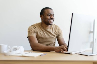 Masaüstü bilgisayarında çalışan gülümseyen Afrikalı Amerikalı adam. Etrafı temiz ve minimalist çalışma alanıyla çevrili yakışıklı siyah erkek. Masasında beyaz bardak ve kulaklıklar var.