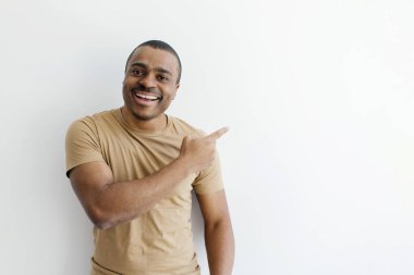 Gülümseyen Afro-Amerikalı adam hevesle kenarı işaret ediyor, bej tişört ve kot giyen adam stüdyo ortamında temiz beyaz arka plana karşı duruyor, boşluğu kopyalıyor.