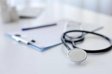 Arka planda tıbbi not panosu ve kalemle beyaz masanın üzerinde duran steteskop yakın plan. Olay yeri parlak ve minimal, temiz ve düzenli bir tıbbi çalışma alanını temsil ediyor..