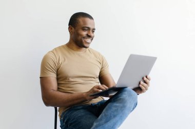 Neşeli siyahi adam dizüstü bilgisayar kullanıyor, beyaz arka planda otururken internette çalışıyor, Afrikalı Amerikalı adam internette internete bakıyor ya da web sitesinden alışveriş yapıyor,