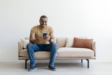 Gülümseyen genç siyah adam rahat rahat koltukta dinleniyor ve akıllı telefon kullanıyor, yakışıklı Afro-Amerikan erkek cep telefonuyla oturma odasında dinleniyor, mesajlaşıyor ya da internette geziniyor.