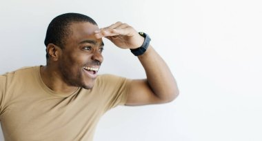 Gülümseyen siyahi adam, elini alnına yaklaştırıyor ve ilgiyle fotokopi uzayına bakıyor, neşeli Afrikalı Amerikalı yakışıklı erkek, beyaz arka planda dikilirken ilginç bir teklif sunuyor.