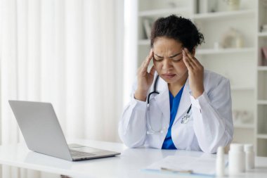 Üniformalı, stresli kadın doktor portresi, iş yerinde baş ağrısı, aşırı çalışan, dizüstü bilgisayarlı ve şakaklarına dokunan, akut migreni olan hasta doktor kadın.
