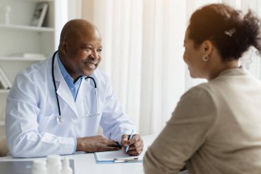 Klinikteki toplantı sırasında üniformalı siyah doktor kadın hastaya danışmanlık yapıyor, beyaz önlüklü ve stetoskop takan terapist, masada oturan ve genç kadını dinleyen...