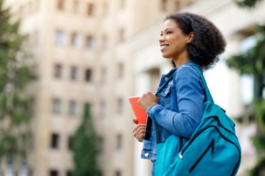Sırt çantası ve not defteri olan Afro-Amerikalı genç bir kadının yandan görünüşü, gülümseyen siyah bir kadın, üniversite binasının yanında neşeli bir ifadeyle yürürken,