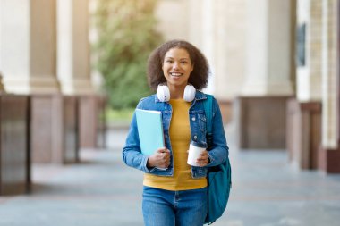 Kahve fincanı ve çalışma kitaplarıyla yürüyen gülümseyen siyah öğrenci, kulaklık ve sırt çantası takan mutlu Afro-Amerikan kadın şehir koleji ortamında kameraya bakıyor.