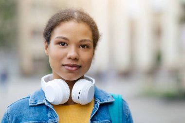 Boynuna kulaklık takmış Afro-Amerikalı genç bir kadının portresi. Güzel, siyah bir kadın, yumuşak bir arka plan bulanıklığıyla kameraya özgüvenle bakıyor.