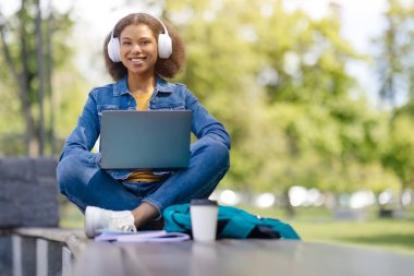 Kablosuz kulaklıkla gülümseyen siyah kız öğrenci dışarıda dizüstü bilgisayarla çalışıyor, genç Afro-Amerikan kadın kampüste bankta bilgisayarla oturuyor, uzaktan öğrenmenin keyfini çıkarıyor,