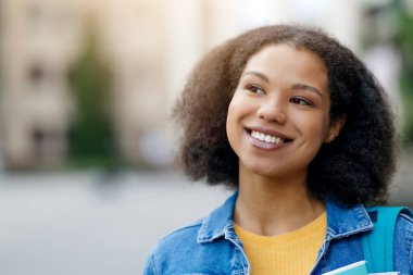 Yan tarafa bakan mutlu siyahi kadın öğrenci portresi, güzel Afro-Amerikan kadın açık havada, doğal ışık altında gülümsüyor ve rahatlıyor.