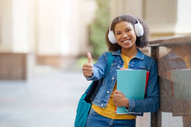 Üniversite binasının yanında ayakta dikilen Güzel Öğrenci Kadının Portresi, Kulaklıklı Gülümseyen Genç Kadın Çalışma kitaplarını tutuyor, Yurtdışında Çalışmayı Öneriyor