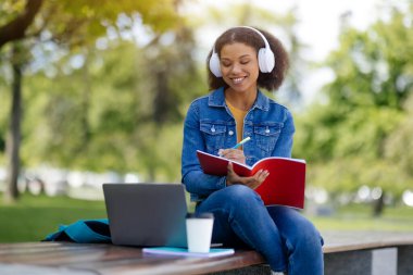 Odaklanmış, beyaz kulaklıklı genç Afro-Amerikalı bir kadın, dışarıda bankta çalışırken kırmızı deftere notlar yazıyor, gülümseyen siyah bir kadın, uzaktan çalışmanın tadını çıkarıyor, boşluğu kopyalıyor.