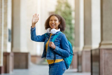 Sırt çantalı ve kulaklıklı neşeli siyahi kadın kampüs girişinde poz veren Afro-Amerikan kadın kameraya merhaba diyor.