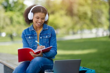 Çevrimiçi eğitim, uzaktan öğrenme konsepti. Genç siyahi kadın parkta bankta otururken Webinar 'a katılıyor, Afro-Amerikan kadın dizüstü bilgisayar ve kulaklık kullanıyor, notlar alıyor, yer kopyalıyor.