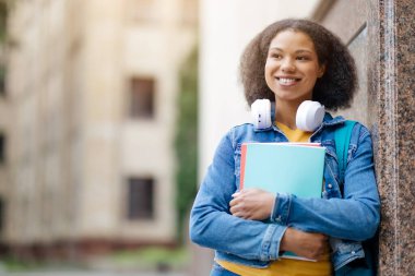 Ellerinde çalışma kitaplarıyla, duvara yaslanmış, kolları çaprazlanmış, kulaklıkları boynunda kulaklıkları olan Afro-Amerikan bir kadın öğrenci fotokopi odasına bakıyor.