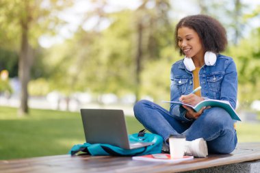 Afrika kökenli Amerikalı kız öğrenci ders kitabına not alıyor ve kampüs parkında otururken dizüstü bilgisayar kullanıyor, gülümseyen siyahi kadın uzaktan çalışıyor, online eğitimin tadını çıkarıyor,