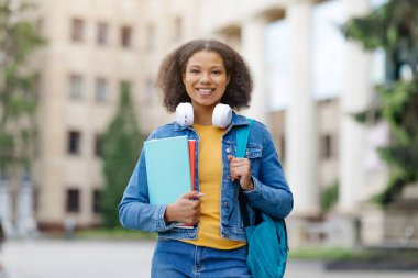 Sırt çantası ve defterleriyle dışarıda duran gülümseyen siyah kız öğrenci, kulaklık takan mutlu Afro-Amerikan kadın, kampüste rahat ve kendinden emin hisseden,