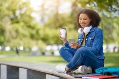 Parkta bacak bacak üstüne atmış bir şekilde oturan Afro-Amerikan öğrenci elinde akıllı telefon ve kahve fincanı, boynunda dinlenen kulaklıklar, fotokopi çekerken gülümsüyor.