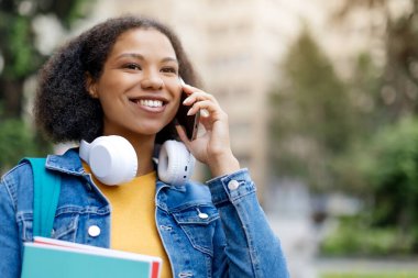 Neşeli Afrikalı Amerikalı kız öğrencinin cep telefonuyla konuşan, kampüste çalışma kitabıyla gezen, kenara bakan ve akıllı telefon aracılığıyla iletişim kuran yakın plan portresi. Mobil İletişim