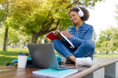 Mesafe öğrenme. Genç siyah kız öğrenci dışarıda dizüstü bilgisayarla çalışıyor bilgisayarını kullanan mutlu Afrikalı Amerikalı kadın ve parkta bankta otururken not defteri tutuyor.