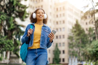 Afro-Amerikalı neşeli genç bir bayan kampüste akıllı telefon ve kulaklıkla yürüyor, mutlu siyah kadın favori müziği dinliyor, neşeli ve odaklanmış görünüyor, boşluğu kopyalıyor.