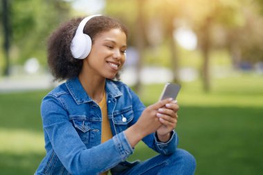 Parkta dinlenirken, müzik dinleyen ve internette gezinen gülümseyen siyahi kız öğrenci, kulaklık takan mutlu Afro-Amerikan kadın en sevdiği müzik listesinin tadını çıkarıyor.