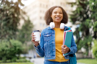 Bir elinde kahve fincanı, diğer elinde kitaplarıyla dışarıda gülümseyen, neşeli, siyah bir kadın üniversite avlusunda kameraya bakıyor, boşluğu kopyalıyor.