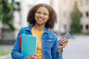 Gülümseyen Afro-Amerikan öğrenci kadın çalışma kitapları ve akıllı telefon tutuyor, arka planda kampüste mutlu siyah kadın duruyor, rahat ve neşeli bir ifade, serbest alan.