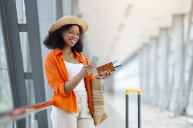 Havaalanı salonunda beklerken cep telefonunda gezinen siyahi bir kadın, Afro-Amerikan bir kadın seyahat uygulaması kullanıyor veya uçuş bilgilerini kontrol ediyor.
