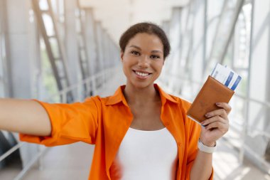 Güzel siyah kadın selfie çekiyor, uçuş biletleri ve pasaportu var, mutlu Afro-Amerikan kadın havaalanı terminalinde duruyor, tatilde eğlenen neşeli bayan.