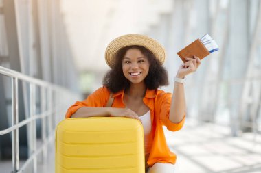 Yolculuk için hazır. Bavul ve uçuş biletleriyle poz veren mutlu siyah kadın, saman şapkalı neşeli Afro-Amerikan kadın havaalanında beklerken kameraya gülümsüyor.