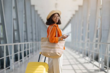 Saman çantalı, sarı bavullu şık siyah kadın omzunun üzerinden bakıyor, mutlu Afro-Amerikan kadın elinde telefon, kameraya gülümsüyor, uzay kopyaları yapıyor.