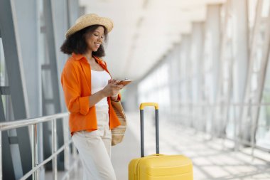 Havalimanında beklerken akıllı telefon kullanan güzel siyahi kadının portresi, Terminalde bavulla dururken gülümseyen Afro-Amerikan kadın cep telefonu uygulaması,