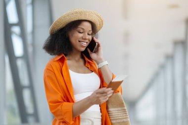 Havalimanında bekleyen güzel siyah kadın cep telefonuyla konuşuyor, Milenyum Afrikalı Amerikalı bayan biniş kartına bakıyor ve keyifli cep telefonu sohbetinin tadını çıkarıyor,