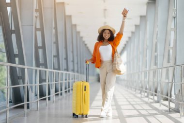 Sonunda tatil. Mutlu, heyecanlı, Afro-Amerikan kadın havaalanında sarı bavulla poz veriyor, neşeli siyah kadın elini kaldırıyor ve gülümsüyor, hasır şapka giyiyor, fotokopi odası, boy boy