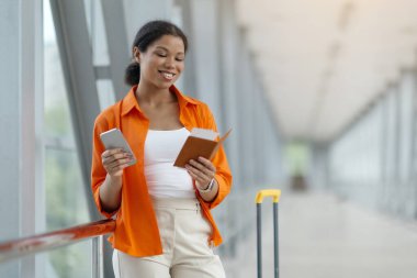 Pasaport okurken gülümseyen ve parlak havaalanı koridorunda akıllı telefon kullanan siyahi bir kadın, bavuluyla bekleyen mutlu bir Afro-Amerikan kadını, seyahat belgelerini kontrol eden,