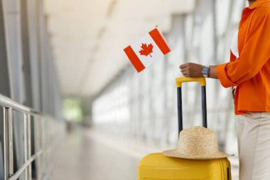 Havaalanında elinde Kanada bayrağı, bavulu ve hasır şapkası olan tanınmamış Afrikalı Amerikalı bir kadın Kanada 'ya seyahat etmeyi sembolize ediyor.