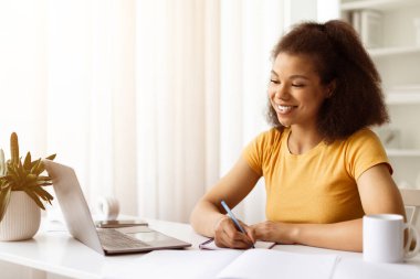 Gülümseyen Afrikalı Amerikalı kadın dizüstü bilgisayarla evde çalışırken deftere yazı yazıyor, genç siyahi kadın online bir derse veya toplantıya katılıyor, meşgul ve odaklanmış görünüyor, boşluğu kopyalıyor.