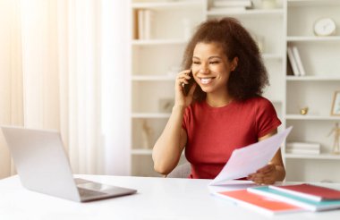 Cep telefonuyla konuşan ve elinde kağıtla dizüstü bilgisayarla oturan gülümseyen siyahi kadın, İçişleri Bakanlığı 'nda çalışan Afro-Amerikan kadın. Mesafe Serbest Görev Konsepti