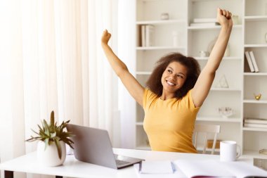 Gülümseyen siyahi kadın serbest çalışan, ofisteki çalışma gününden sonra gerinen, Afro-Amerikan kadın, dizüstü bilgisayarla masada oturuyor, çevrimiçi projeyi bitirdikten sonra dinleniyor, uzayı kopyalıyor.