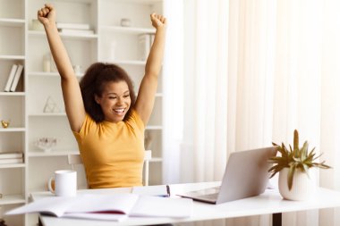 Genç siyahi bir kadın, dizüstü bilgisayarla masasında otururken ellerini havaya kaldırıyor. Afrikalı Amerikalı bir kadın başarıyı kutluyor. Çevrimiçi çalışmanın ya da çalışmanın olumlu sonuçlarından keyif alıyor.