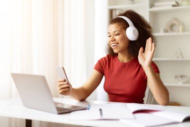 Afro-Amerikalı kız öğrenci akıllı telefon ve dizüstü bilgisayar kullanarak neşeli bir şekilde el sallıyor, mutlu siyah kadın evde rahat rahat otururken sanal sohbete dalıyor.