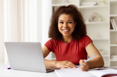 Masasında dizüstü bilgisayarla oturan neşeli siyahi kadın portresi, mutlu Afro-Amerikan bayan öğrenci bilgisayarla çalışıyor ve notlar alıyor, kameraya bakıyor ve gülümsüyor.