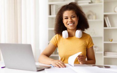 Evdeki masada çalışan mutlu zenci kız öğrenci portresi, dersten sonra ara veren, boynunda kulaklıkları olan neşeli Afro-Amerikan kadın. Kameraya içten bir şekilde gülümsüyor.