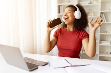 Neşeli Afro-Amerikan öğrenci kadın akıllı telefona şarkı söylüyor, kulaklık takıyor ve evde eğleniyor, neşeli siyah kadın dizüstü bilgisayarla masada otururken eğleniyor,